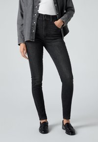 Personne portant un jean skinny noir taille haute, une ceinture noire, une chemise en jean noire boutonnée par-dessus un haut blanc, et des mocassins noirs, debout avec une main dans la poche.