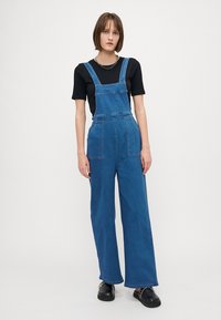 Denim kombinezon v svetlo modri barvi z širokimi hlačnicami in prilagodljivimi naramnicami. V kombinaciji s črno majico s kratkimi rokavi in črnimi natikači.