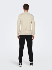 Helle beige Strickjacke mit langen Ärmeln, rundem Ausschnitt und tailliertem Design. Kombiniert mit schwarzen Hosen und blau-weißen Sneakers.