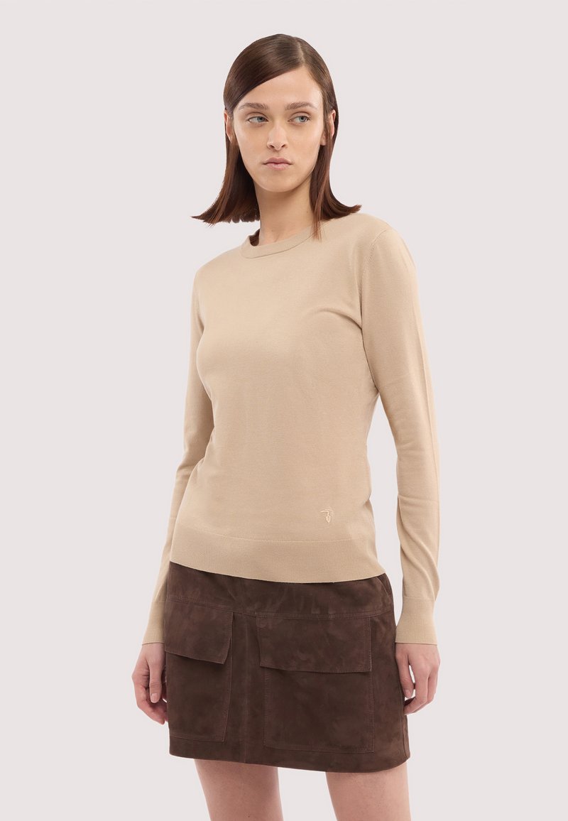 Junge Frau mit glatten braunen Haaren, die einen beigefarbenen Pullover mit langen Ärmeln und einen dunkelbraunen Minirock aus Veloursleder mit Taschen trägt.