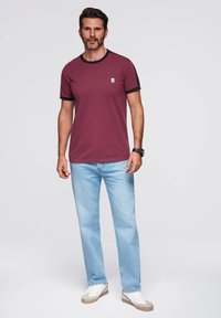 Burgundské tričko s černými detaily, spárované se světle modrými džíny a bílými teniskami. Mezi významné detaily patří malá logo nášivka na tričku.