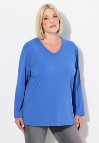 Maglietta blu a maniche lunghe con scollo a V, realizzata in tessuto morbido, con una vestibilità rilassata e orlo dritto, abbinata a jeans grigi. Design semplice e versatile.
