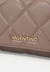 Αυτός ο καπιτονέ καφέ δερμάτινος σάκος διαθέτει το χρυσό λογότυπο "Valentino". Έχει υφή με διαγώνια ραφή και έντονη λεπτομέρεια στην επωνυμία.