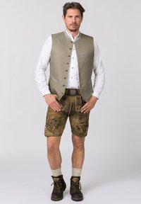 Tweed-Weste in olivgrün, darunter ein weißes Hemd, braune Lederhosen mit eingeprägten Blumenmustern und dunkle Schnürstiefel.