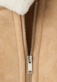 Beige Wildlederkleidung mit einem sichtbaren metallischen Reißverschluss. Der Reißverschluss hat eine glänzende Oberfläche und verläuft vertikal entlang der Vorderseite, mit sichtbaren Stichen.