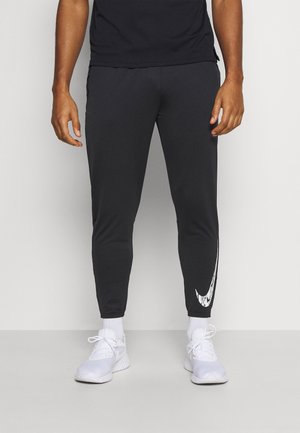 Man die zwarte sportieve joggers draagt met wit swoosh-logo op het linkerbeen, witte sokken en witte sneakers, staand tegen een neutrale muur.