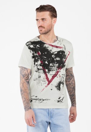 Mann mit Tätowierungen an den Armen trägt ein weißes T-Shirt mit großem schwarzem Schädeldesign und roten Dreieck, kombiniert mit hellblauen Jeans, blickt nach links.