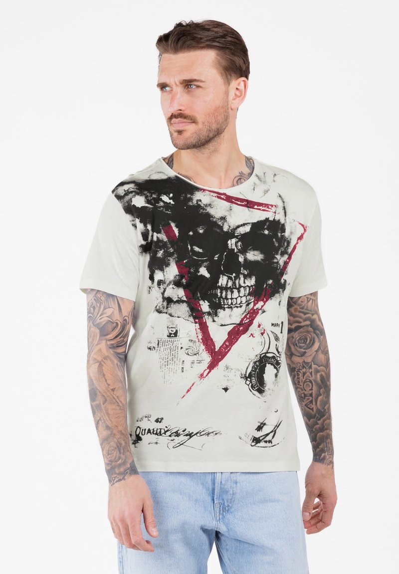 Man met tatoeages op zijn armen draagt een wit T-shirt met een grote zwarte schedel en een rode driehoek graphic, gecombineerd met lichtblauwe spijkerbroek, kijkt naar links.