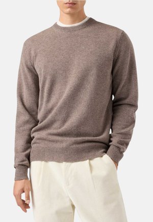 Uomo che indossa un maglione marrone chiaro a maniche lunghe sopra una camicia bianca con pantaloni a coste color crema, mani rilassate lungo i fianchi.