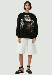 Schwarzer Hoodie mit Blumendruck und dem Text "ALLSAINTS LONDON", kombiniert mit weißen Shorts und schwarzen Sandalen mit Riemchen.