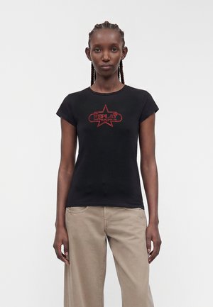 Slank kvinde med flettet hår iført sort tætsiddende T-shirt med rødt stjerne-logo og beige bukser med vide ben, står mod en ensfarvet baggrund.
