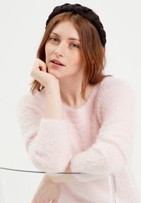 Zwarte gevlochten haarband, zachtroze fuzzy trui met een ronde halslijn en geribbelde manchetten, model steunt met de kin op de hand tegen een heldere stoel.