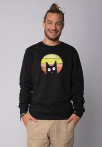 Zwarte sweatshirt van katoen met een ronde afbeelding van een zwarte kat tegen een zonsondergangsverloop van geel, oranje en rood.