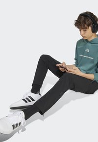 Ungdom som bär en teal hoodie med vit logotyp, svarta byxor och vita skor med svarta ränder, sittande och använder en smartphone. Svarta hörlurar.