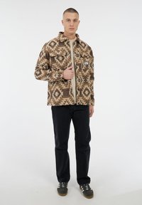 Man staat in een bruine jack met geometrisch patroon, een beige shirt, zwarte broek en grijze sneakers op een effen witte achtergrond.