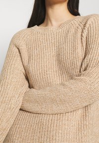 Femme portant un pull beige en tricot avec un col rond, montrée du cou à la taille, les bras croisés sur la poitrine.