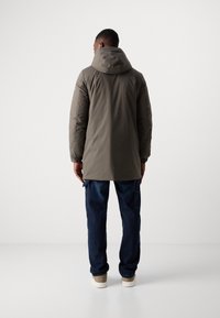 Padded parka in dark grey with a hood, long design, elastic sleeves, paired with dark blue jeans and beige sneakers, viewed from the back.