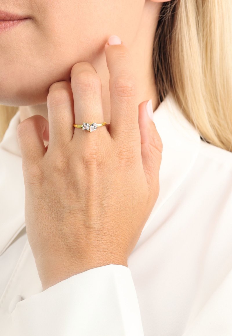 Złoty pierścionek z dwoma przezroczystymi, trójkątnymi kamieniami szlachetnymi. Obrączka jest cienka i wypolerowana, podkreślająca blask kamieni. Dłoń oparta na twarzy.