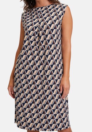 Mouwloze jurk tot op de knie met een geometrisch zigzagpatroon in marineblauw, beige en wit, gedragen door een vrouw die voor een effen achtergrond staat.