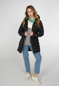Sort polstret frakke med hætte, med lynlåslukning foran og sidelommer, lagd over en grå top med en grøn halsvarmer og blå jeans.