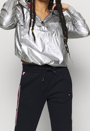 Osoba nosząca błyszczącą srebrną kurtkę przeciwwiatrową i czarne spodnie dresowe z czerwonymi i białymi paskami z boku, poprawiająca włosy.