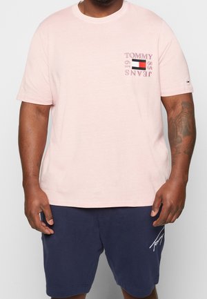 Mann trägt ein hellrosa Tommy Jeans T-Shirt mit Logo auf der Brust und marineblaue Shorts mit charakteristischer Stickerei am linken Bein.