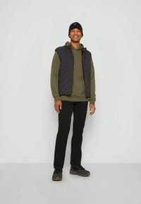 Čierna nafukovacia vesta na olivovo zelenej mikine, v kombinácii s čiernymi nohavicami a tmavými topánkami. Vesta má prešívaný dizajn a kapucňu.