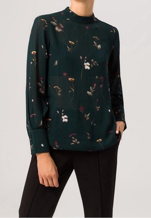 Long-sleeve dark green blouse with small floral print worn with black pants, one hand resting in pants pocket.