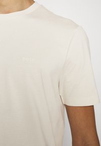 T-shirt beige clair avec un col rond, des manches courtes et un logo "BOSS" en relief subtil sur la poitrine. Texture du tissu douce et lisse.