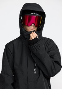 Veste noire imperméable avec une capuche, comprenant une poche zippée et des poignets élastiques, assortie à de grandes lunettes de ski colorées.