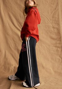 Felpa rossa con cappuccio e pantaloni della tuta neri con lettering rosa e righe bianche ai lati. L'outfit presenta una vestibilità comoda e un tessuto morbido.