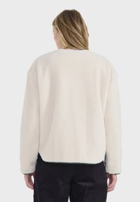 Chaqueta de forro polar color crema con una superficie texturizada y hombros caídos, que presenta un escote redondo y un dobladillo oscuro en contraste.