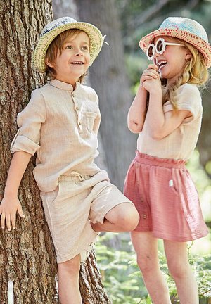 Hellbeige Leinenhemd mit hochgerollten Ärmeln, passende Shorts und einem gewebten Strohhut. Mädchen in einem hellen Oberteil, rosa Knopfrock und Sonnenhut.