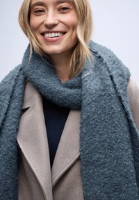 Mørk teal, fluffy tørklæde lavet af tekstureret stof, draperet omkring en lysebeige frakke, båret over en marineblå top.