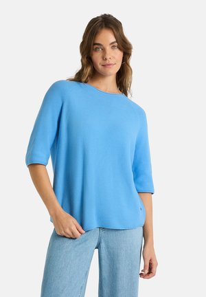 Frau mit welligem braunem Haar trägt einen hellblauen Pullover mit halblangen Ärmeln und hellblaue Jeans vor einem schlichten weißen Hintergrund.