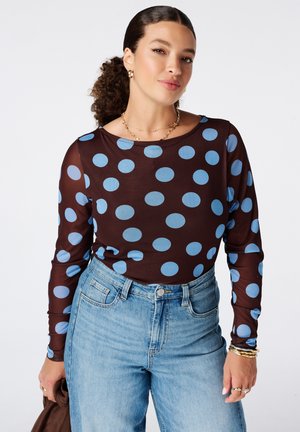 Femme aux cheveux bouclés en queue de cheval portant un haut foncé à gros pois bleus et un jean bleu clair taille haute, tenant une veste marron.