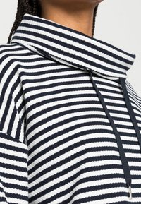 Pruhovaný pulovr s vysokým límcem v námořnické modři a bílé barvě. Disponuje texturovanou látkou, stahovací šňůrkou a jemným leskem kovových částí.