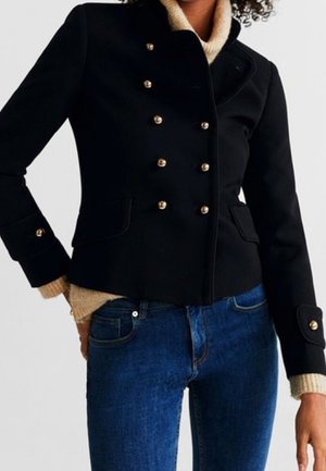Kvinde iført en tætsiddende sort dobbeltradet jakke med guldknapper over en beige rullekravesweater og blå jeans.