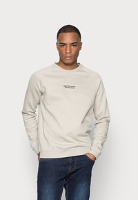 Felpa leggera color beige con scollatura a girocollo e maniche raglan. Presenta un testo minimalista nero che dice "COSTRUZIONI ARTIGIANALI PER UN DENIM".