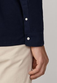 Granatowa koszula z długim rękawem z guzikami przy mankietach, wykonana z materiału bawełnianego o gładkiej teksturze, w połączeniu z jasnymi spodniami.