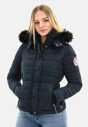 Jeune femme portant une veste matelassée bleu marine avec une capuche en fourrure noire et un jean bleu clair, les mains dans les poches, sur fond blanc.