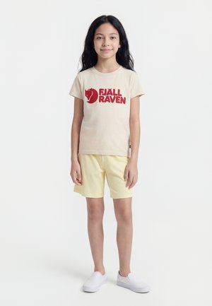 Girl standing, wearing beige Fjällräven t-shirt with red logo, light yellow shorts, and white slip-on shoes on plain white background.