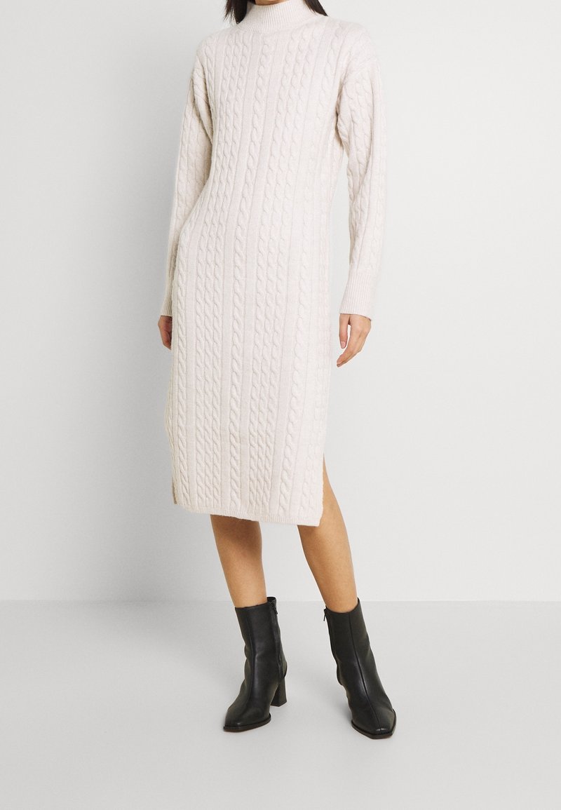 Robe-pull en maille torsadée beige clair, avec un col montant, des manches longues, des fentes latérales et un ourlet mi-long. Bottines noires à talons.