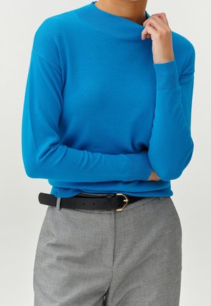 Pull à col roulé bleu côtelé avec des manches longues, associé à un pantalon gris et une ceinture noire avec une boucle dorée. Texture lisse.