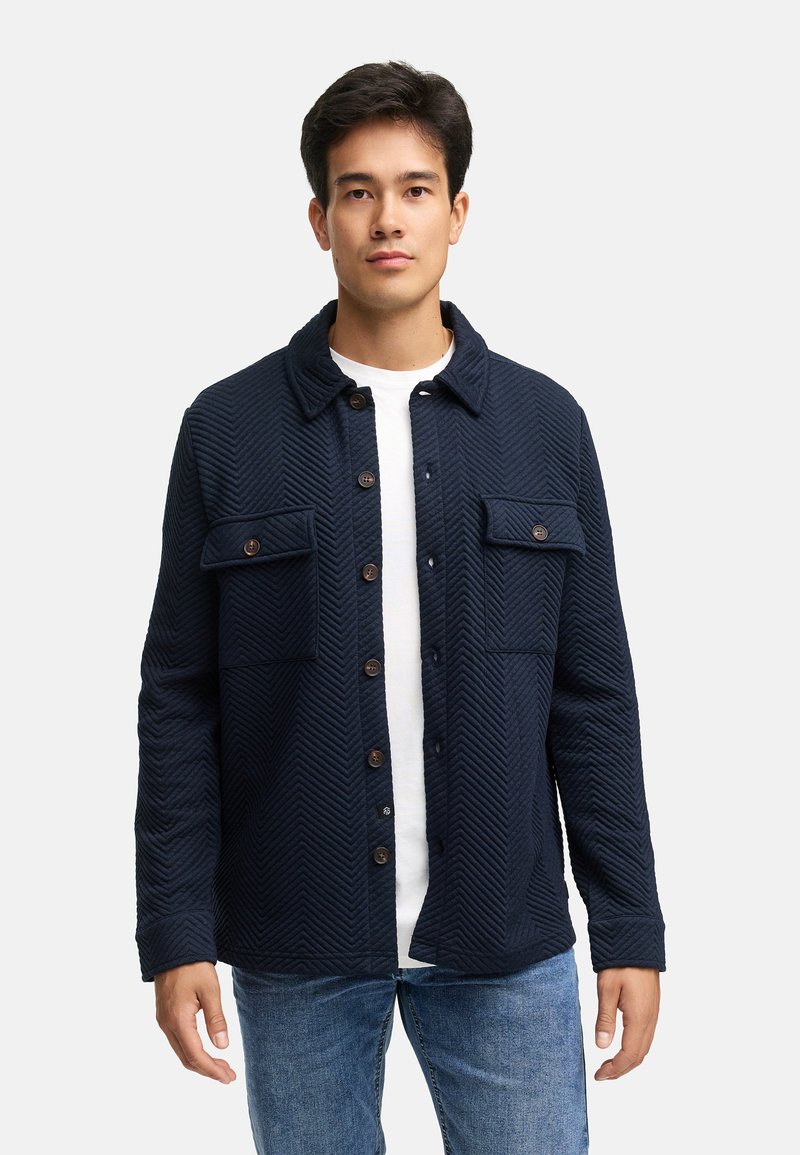 Marineblauer Fischgrätjacke mit Knopfreihe, zwei Brusttaschen und strukturiertem Kragen, aus strukturiertem Stoff. Getragen über einem weißen Hemd.