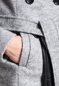 Handen i fickan på en texturerad ljusgrå ullkappa med svarta knappar och ett tygbälte knutet vid midjan.