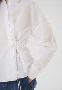 White cotton shirt with a relaxed fit, featuring a tie at the waist and long sleeves. The fabric appears smooth with a crisp design.