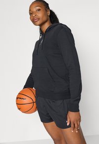 Kvinde i sort hættetrøje og shorts, der holder en orange basketball og står foran en ensfarvet hvid baggrund.