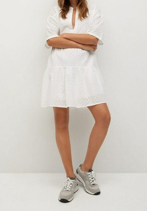 Bílé krátké šaty s dírkovaným vzorem, výstřihem do V a rozšířenou sukní. Nošeny s šedými teniskami s bílými tkaničkami a černými detaily.