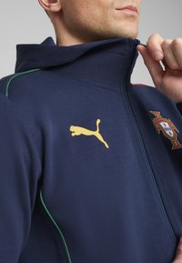 Jūrų laivyno zip-up striukė su žaliais akcentais. Priekyje yra auksinė Puma emblema ir Portugalijos herbas. Pagaminta iš švelnios medžiagos.